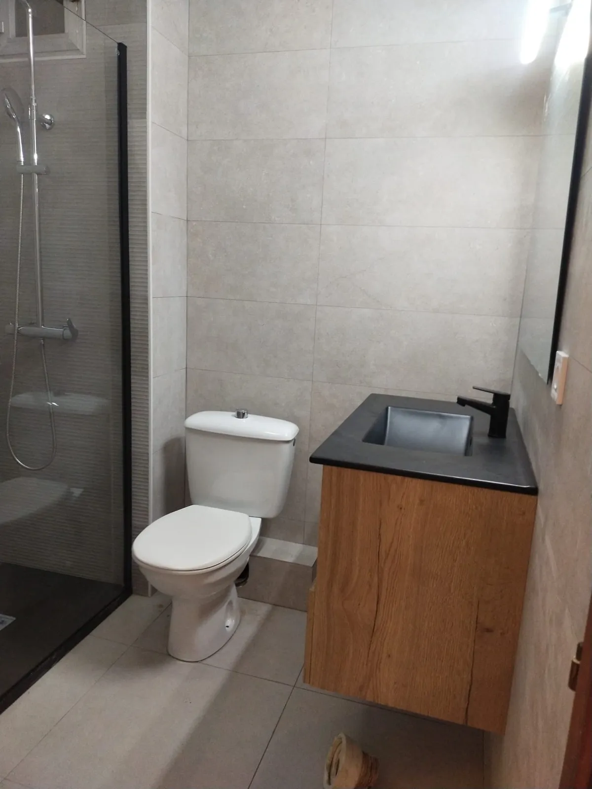 Baño terminado con mampara de perfil negro, mueble de madera, lavabo negro y alicatado gris claro